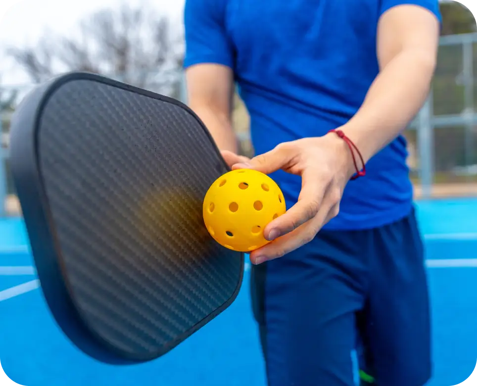 Young adult standing on a blue court, preparing to serve a yellow pickleball with a black paddle, enjoying the dynamic sport during practice or a game