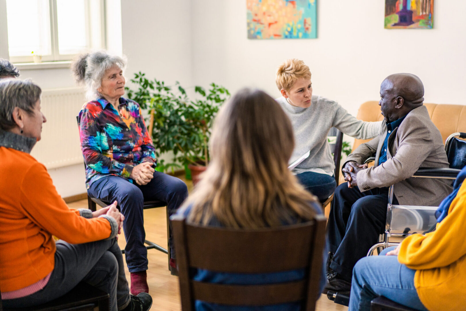 A female psychoanalyst placing a comforting hand onto a mature man's shoulder while conducting a therapy meeting in a bright and airy room with senior residents of a aged care home