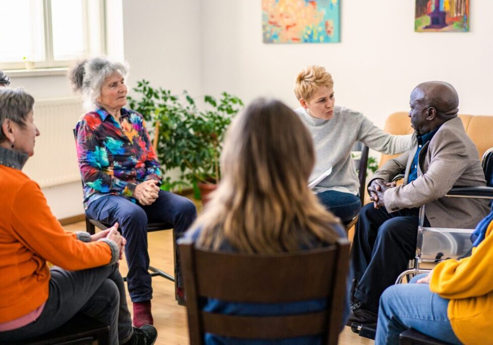 A female psychoanalyst placing a comforting hand onto a mature man's shoulder while conducting a therapy meeting in a bright and airy room with senior residents of a aged care home