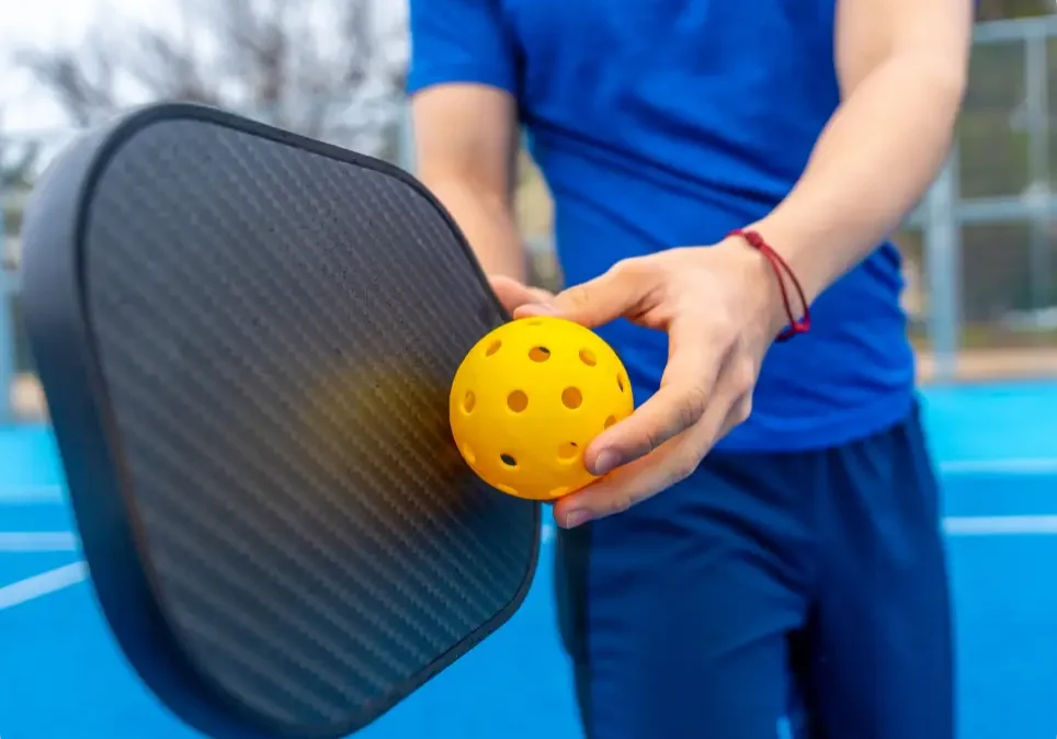 Young adult standing on a blue court, preparing to serve a yellow pickleball with a black paddle, enjoying the dynamic sport during practice or a game